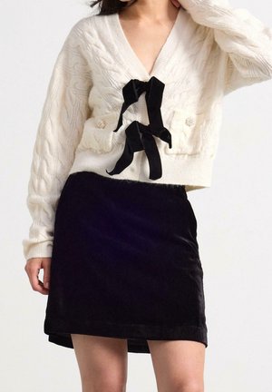 Femme portant un cardigan blanc en maille torsadée avec des liens en ruban noir et des boutons en perles, associé à une mini-jupe en velours noir.