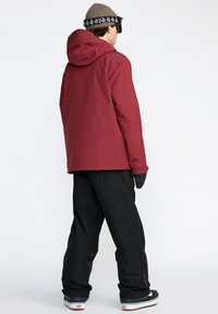 Veste imperméable rouge avec une capuche, portée sur un pantalon de ski noir. Le modèle porte des gants noirs et un bonnet beige avec des accents noirs.