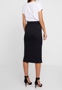 Jupe crayon noire avec une texture côtelée, mettant en avant une silhouette ajustée, des passants de ceinture et un design longueur cheville, associée à des chaussures noires à talons hauts.