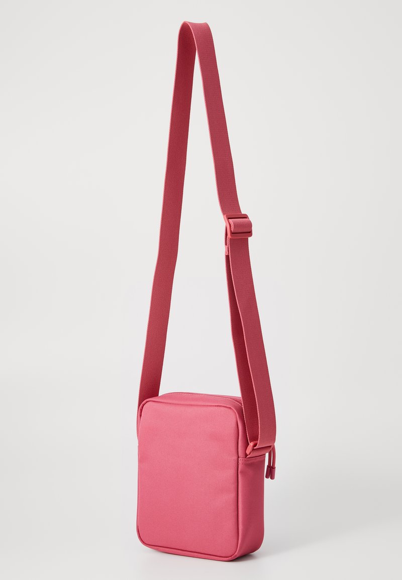 Sac en bandoulière rose en tissu avec une surface texturée, de forme carrée et une sangle réglable. Il dispose d'une fermeture zippée et d'un design minimaliste.