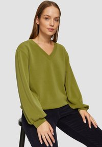 Olijfgroene V-hals sweatshirt met lange, opgeblazen mouwen; zachte textuur; gecombineerd met donkere corduroy broek. Neutrale achtergrond.
