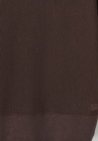 Pull en maille côtelée marron avec une surface texturée et une bande lisse contrastante en bas. Petit logo près de l'ourlet.