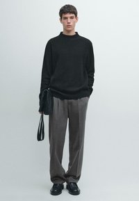 Schwarzer Strickpullover, graue plissierte Hose und schwarze Schuhe. Das Model hält eine kleine schwarze Tasche mit Schulterriemen. Minimalistisches Design und Farben.