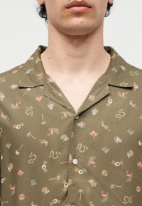 Gros plan d'une personne portant une chemise boutonnée vert olive avec col, ornée de motifs de crânes, de flamants roses, de serpents et de palmiers.