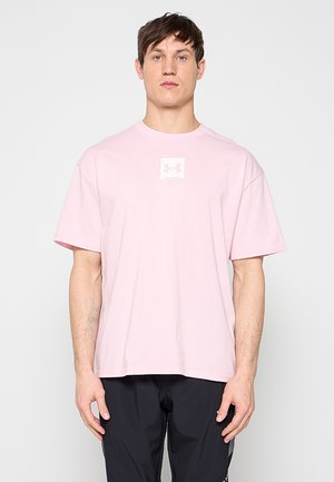 Mladý muž nosící světle růžové triko Under Armour a černé kalhoty stojí proti jednobarevnému bílému pozadí.