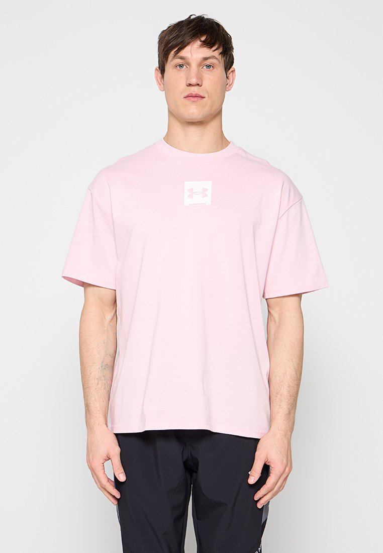 Jeune homme portant un t-shirt Under Armour rose clair et un pantalon noir, debout devant un fond blanc uni.