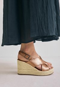 Bronzen leren wedge sandalen met een kruislings strap ontwerp. Beschikt over een geweven jute platform en een verstelbare enkelband met gespsluiting.