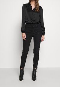 Černá saténová košile na knoflíky s dlouhými rukávy a fitted černé džíny zdobené malými stříbrnými cvočky, doplněné černými kotníkovými botami.