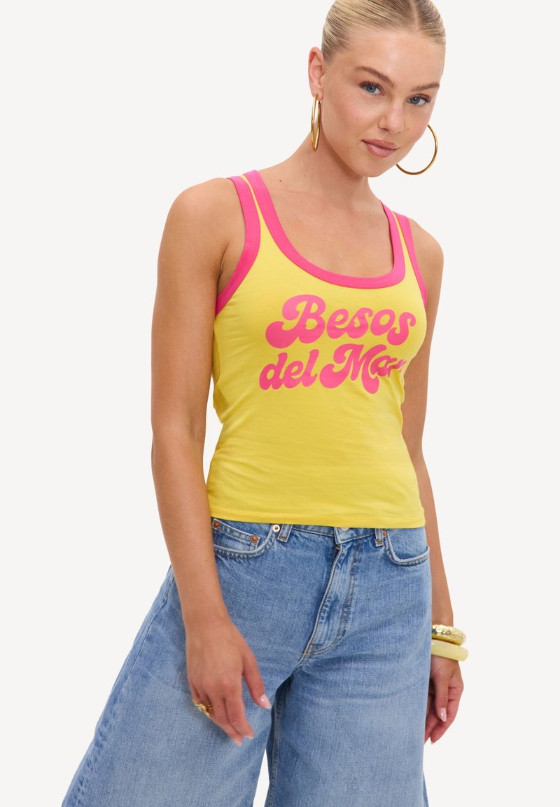 Femme portant un débardeur jaune avec un liseré rose et le texte « "Besos del Mar" », un jean bleu, de grandes boucles d’oreilles créoles dorées et des bracelets au poignet.