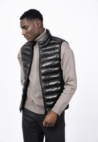 Mustavaha pufferliivi, jossa on tikatua tekstuuria, korkea kaulus, vetoketjusulku, käytetty beige pyllypuseron ja harmaiden housujen päällä. Yksinkertainen muotoilu.