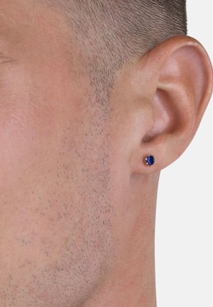Nahaufnahme eines Ohres eines Mannes mit kurzen Haaren, der einen kleinen runden blauen Edelsteinstecker am Ohrläppchen trägt.