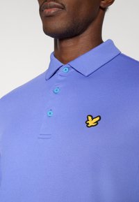 Vijolična polo majica iz teksturirane tkanine z nizkim ovratnikom in svetlo modrimi gumbi. Na prsih ima črno-rumen logotip ptice.