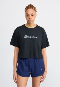 Juoda trumpa maikė su balta "Nike Sportswear" logotipu, derinama su tamsiai mėlynais raštuotais šortais, kuriuose yra mažas baltas "Nike" swoosh logotipas.