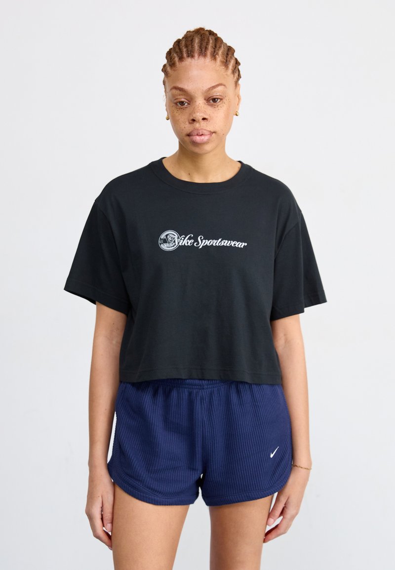 Juoda trumpa maikė su balta "Nike Sportswear" logotipu, derinama su tamsiai mėlynais raštuotais šortais, kuriuose yra mažas baltas "Nike" swoosh logotipas.