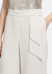 Biały kombinezon z cekinowym topem, szeroką nogawką, kieszeniami po bokach oraz drapowanym wykończeniem z przodu dla dodatkowej tekstury.