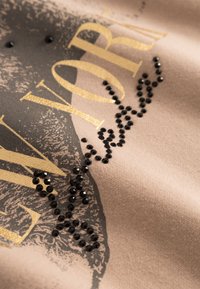 Beżowa tkanina z nadrukowanym wzorem, na którym widnieje napis "NEW YORK" w złotym kolorze, ozdobiony czarnymi rytowanymi kamieniami w zakrzywionym wzorze.