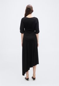 Γυναίκα με μακριά καστανά μαλλιά στέκεται γυρισμένη με την πλάτη, φορώντας μαύρο ασύμμετρο μίντι φόρεμα και μαύρα σανδάλια με τακούνι σε απλό φόντο.