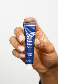 Tube souple bleu marine de "rms Liplights", avec du texte blanc. Le bas présente un capuchon à vis blanc. Tenu dans une main avec du vernis à ongles clair.