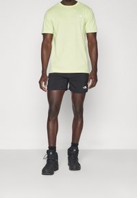 Světle zelené bavlněné tričko, černé kraťasy a černé turistické boty. Tričko má na hrudi malé logo; kraťasy obsahují podobné logo.