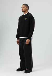 Zwarte trui met geribbelde manchetten, een ronde halslijn en een logo aan de voorkant. Draagt over een wit T-shirt, gecombineerd met losse zwarte spijkerbroek en zwarte laarzen.