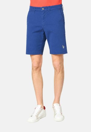 Uomo con pantaloncini blu fino al ginocchio e scarpe da ginnastica bianche con dettagli rossi, in piedi davanti a uno sfondo chiaro e uniforme.