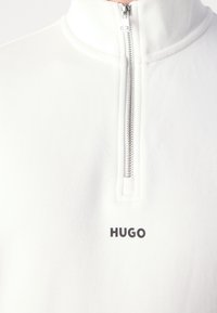 Pullover bianco a zip corta realizzato in tessuto morbido, con una cerniera silver e un piccolo logo nero "HUGO" nella zona del petto.