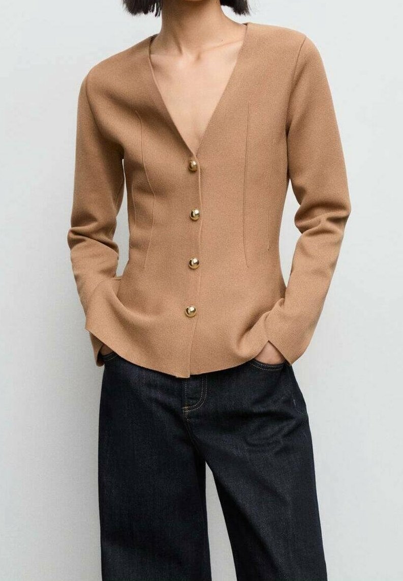 Femme portant un cardigan cintré couleur camel avec des boutons dorés et un jean denim foncé à jambes larges, mains dans les poches, sur un fond uni.