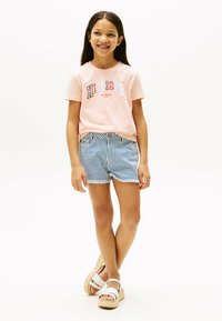 Lyserød bomulds-t-shirt med "HILFIGER" grafik, parret med blå og hvide stribede denimshorts. Hvide platform sandaler fremhæver outfittet.