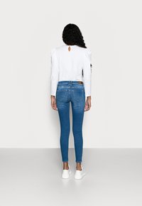Slanke blauwe spijkerbroeken met achterzakken, gedragen met een witte blouse met lange mouwen en een sleutelgatdetail op de rug. Witte sneakers.
