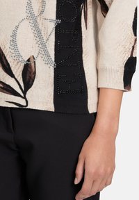 Pull en tricot beige avec motif floral foncé, esperluette en strass, et panneau noir avec lettres en strass sur la manche et le torse.