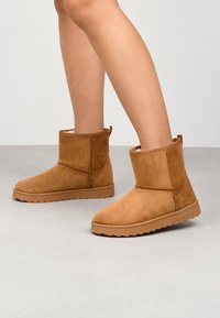 Stivaletti alla caviglia in suede marrone con suola in gomma piatta, punta arrotondata e design con cuciture a vista. Fodera morbida visibile lungo il bordo superiore.