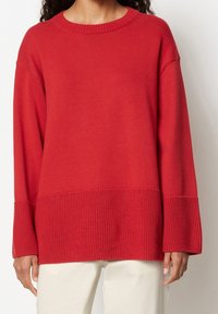 Pull rouge en tricot avec un large ourlet et des poignets côtelés, présentant une coupe décontractée et un col rond. Texture lisse avec un détail de couture visible.