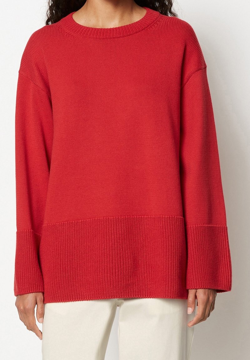 Pull rouge en tricot avec un large ourlet et des poignets côtelés, présentant une coupe décontractée et un col rond. Texture lisse avec un détail de couture visible.