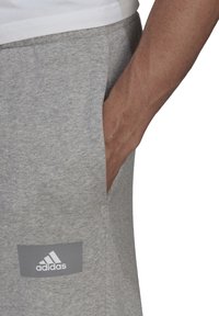 Šedé tepláky vyrobené z bavlněné směsi, s bočními kapsami a šedou nášivkou loga Adidas na levém stehně. Textura vypadá měkce.