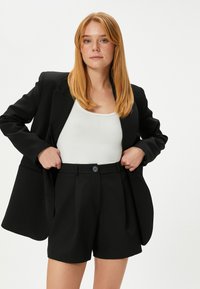 Zwarte getailleerde blazer over een witte geribde tanktop, gecombineerd met zwart hoge taille shorts met knoopsluiting en geplooide voorkant.