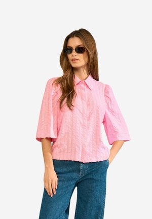 Kobieta nosząca prostokątne czarne okulary przeciwsłoneczne, różową bluzkę w paski z szerokimi rękawami oraz niebieskie dżinsy, stojąca z jedną ręką w kieszeni.