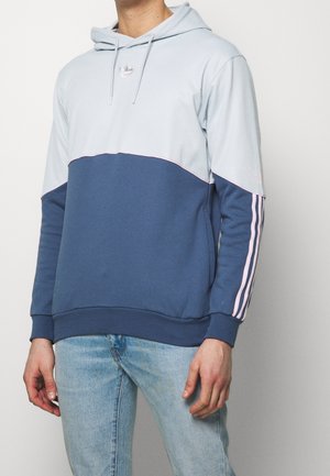 Sweat à capuche en bleu clair et marine avec des accents roses, présentant un design divisé et trois rayures le long des manches. Tissu doux.