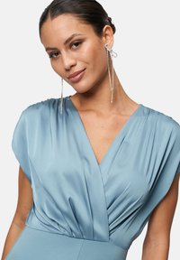 Hellblaues Kleid mit tiefem V-Ausschnitt, weichem, drapiertem Stoff und gefalteten Details am Oberteil. Begleitet von langen, funkelnden Ohrringen.