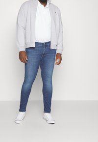 Šedá mikina na zip, bílá polo tričko, modré slim-fit džíny a bílé tenisky s červenými akcenty. Hladký materiál, ležérní design.