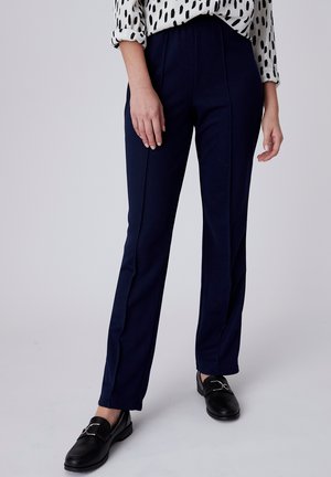 Femme portant un pantalon bleu marine sur mesure, des mocassins noirs et un chemisier blanc à manches longues avec des taches abstraites noires, debout.