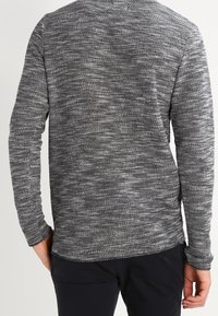 Man som bär en långärmad, grå och vit melerad stickad tröja, sedd bakifrån, tillsammans med svarta byxor.