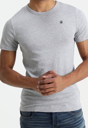 Homme portant un t-shirt gris clair ajusté à manches courtes avec un petit logo noir sur la poitrine, les mains jointes devant, portant un jean bleu foncé.