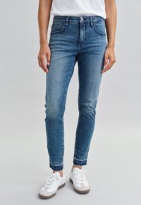 Jeans skinny en denim bleu pour femmes avec une finition délavée, taille mi-haute, design à cinq poches et ourlet effiloché. Portés avec des baskets blanches.