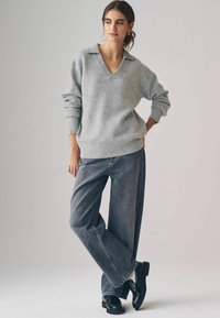 Maglione grigio a maglia con scollo a V e polsini a costine, abbinato a jeans grigi scuri dalla vestibilità ampia e scarpe nere. Design semplice, senza motivi.
