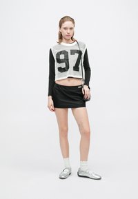 Korte zwarte lange-mouwen top met witte mesh en grote "97" print, gecombineerd met een zwarte miniskirt, zilveren schoenen en witte sokken.