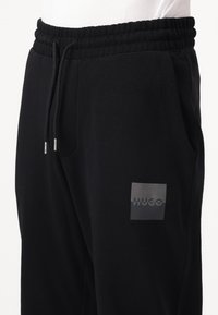 Black sweatpants made of soft fabric, featuring an elastic waistband, side pockets, and a grey logo patch on the thigh.