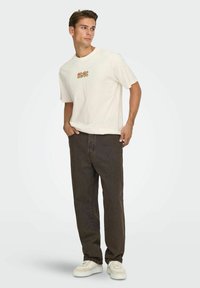 White cotton t-shirt with orange AC/DC logo, paired with dark brown denim pants and white sneakers. Model stands, hands in pockets.