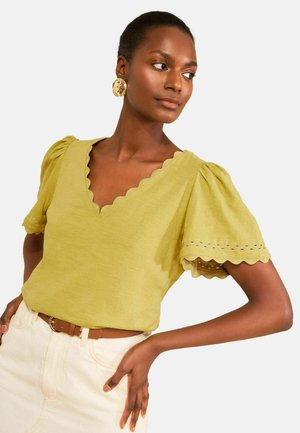 Naine kannab helerohelist, siksakäärisega V-kaelusega pluusi, millel on volangilised varrukad, ja kreemikaid pükse, poseerides käega puusal ja vaadates küljele.