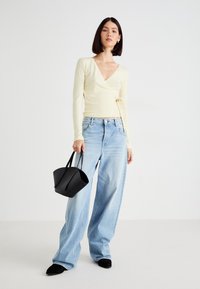 Gul langærmet indpakningsbluse, lyseblå vidde denim jeans, sort håndtaske med korte håndtag og sorte spidse flade sko. Ensfarvet baggrund.