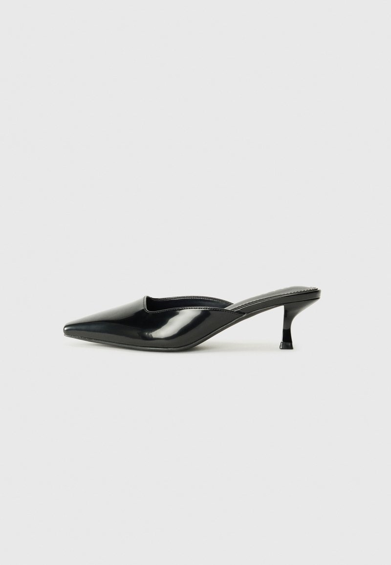 Mule femme en cuir verni noir à bout pointu et petit talon kitten sur un fond clair uni.